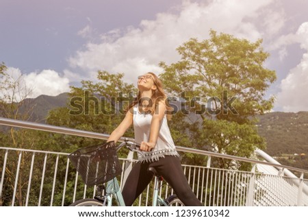 Young attractive woman with bicycle on a bridge surrounded by nature and architecture during a cloudy day