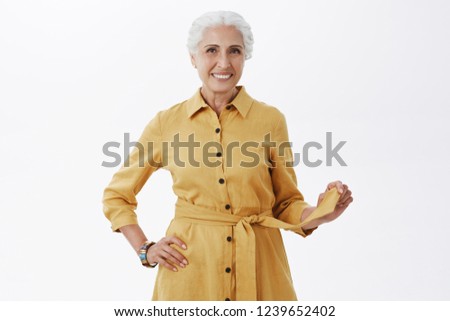 Looking stylish as always. Portrait of energized and charismatic elderly woman in fashionable yellow coat playing with belt and smiling joyfully looking pretty and feeling young over gray background