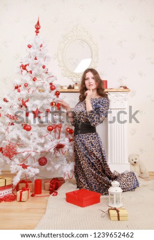 a nice woman in a long leopard dress shyly smiles near a white Christmas tree and a fireplace with gifts