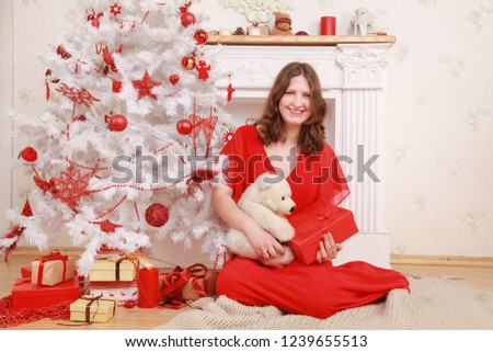 a nice woman in a long red evening dress shyly smiles near a white Christmas tree and a fireplace with gifts