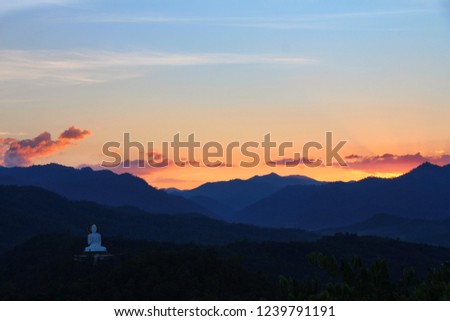 The sunset over the edge of the mountain is light orange, with blue sky, with black clouds floating in ึthe sky. See the white Buddha on the hill to cascade down beautiful.