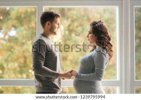 Happy family man and pregnant woman stand in front of big window in their house. They wait for a baby