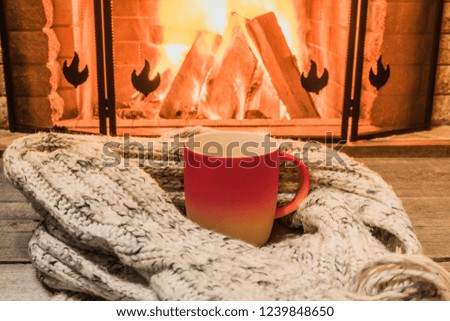Red cup with hot tea and cozy warm scarf near fireplace, in country house, winter vacation, horizontal.