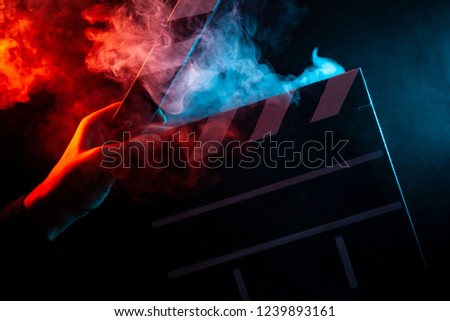Close-up on an open clapper in hand before starting shooting a film with multi-colored smoke around with red and blue backlighting on a black isolated background