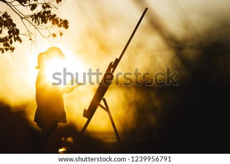 silhouette of a young woman with sun on face painting a picture on canvas on an easel, girl profile with paint brush and palette engaged in art on the nature at sunset