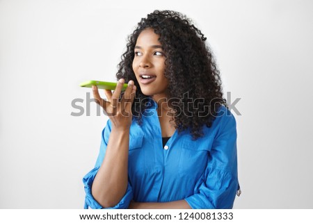 Positive attractive young Afro American female with curly hair holding smart phone, looking away with pensive expression while recording voice message. Modern gadgets, people and communication