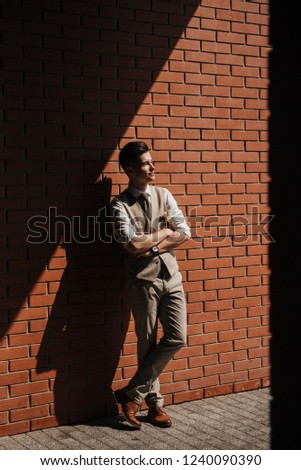 A man in a suit stands against a red brick wall.