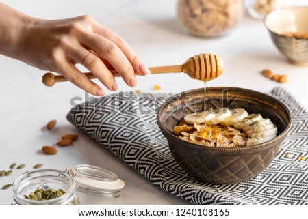 Female hand pours honey into the morning healthy breakfast in a brown bowl with muesli, bananas, sesame seeds and pumpkin. Royalty-Free Stock Photo #1240108165