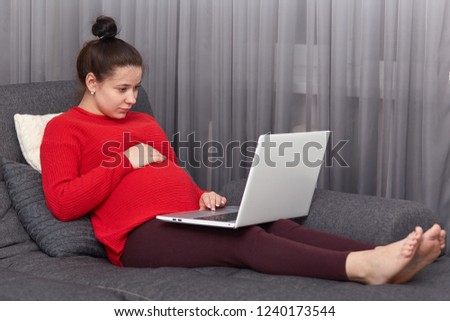 Photo of pregnant woman feels lazy, looks attentively at laptop computer, poses at sofa, watches film online, enjoys free internet connection at home, spends spare time with modern technologies