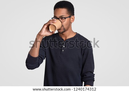 Serious prosperous businessman has coffee break, holds paper cup, enjoys hot drink, wears glasses and casual jumper, stands indoor against white background. People, ethnicity and drinking concept.
