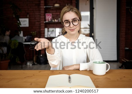 Polite human resources manager in eyewear sitting at her desk extending hand at camera, open to cooperation, making welcoming sign, saying: Please take seat. Friendly businesswoman greeting partner