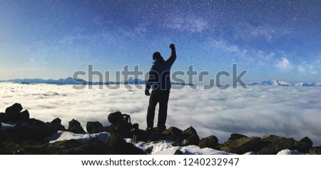 Back view of tourist hiker man with raised arm standing on rocky hill on copy space background of beautiful foggy valley filled with white clouds, snowy mountain tops on horizon and blue starry sky.