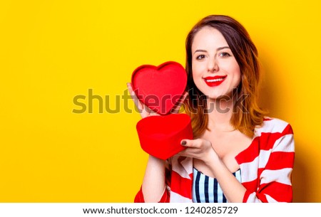 Portrait of beautiful young smiling red-haired white european woman in red striped shirt with heart shape box on yellow background