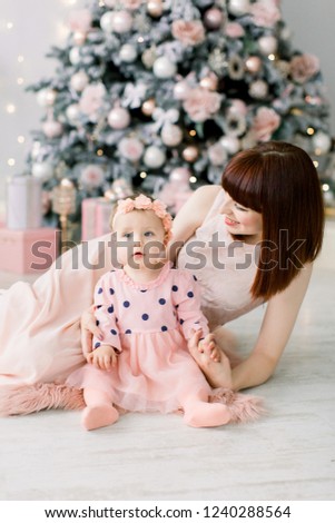 happy mother with her daughter playing near the Christmas tree. Mother kissing and hugging her daughter. Christmas, indoors, cozy room
