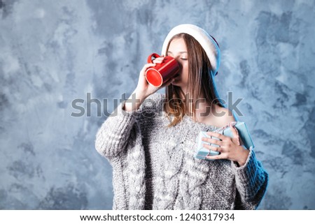 Merry Christmas and happy new year! Adorable happy girl with red cup and blue presents hold in hands. Close portrait on gray background. Girl in santa hat and sweater looks very pleased and drinking