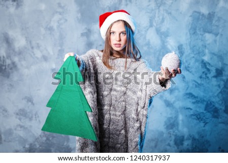 Merry Christmas and happy new year! Adorable female christmas tree and ball in hands. Close portrait on gray background. Girl in santa hat and sweater looks very cheeky