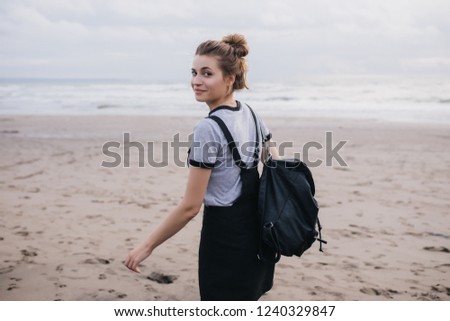 Dreamy girl with black backpack looking over shoulder while walking along coast. Outdoor photo of cheerful white female model spending time at beach.