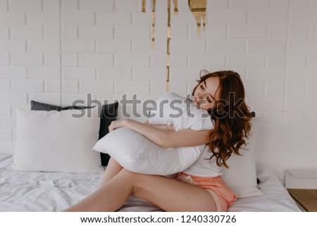 Winsome female model holding white pillow. Indoor photo of carefree red-haired woman sitting on bed.