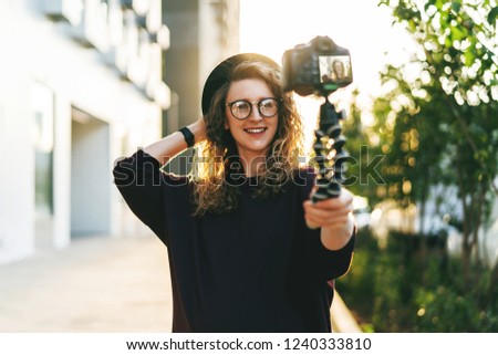 Young female blogger in trendy glasses and hat walks on city street, records herself on video holding camera for tripod. Hipster girl takes pictures of herself on camera. Lifestyle, selfie, backlight.