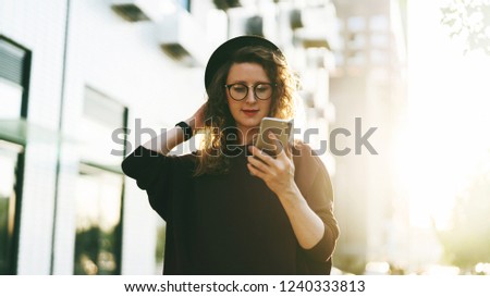 Young curly woman in hat and fashionable glasses makes photo on digital camera of smartphone, standing on city street. Smiling happy woman taking pictures of herself on phone. Sunny day, backlight.