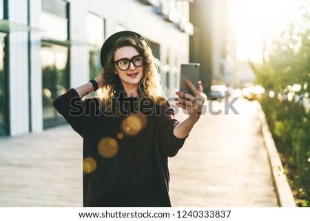 Young curly woman in hat and fashionable glasses makes photo on digital camera of smartphone, standing on city street. Smiling happy woman taking pictures of herself on phone. Sunny day, backlight.