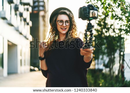 Young female blogger in trendy glasses and hat walks on city street, records herself on video holding camera for tripod. Hipster girl takes pictures of herself on camera. Lifestyle, selfie, backlight.