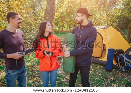 Picture of three friends in forest. Young woman smiles and looks at bearded guy. He holds sleeping bag. Another young man holds knife.