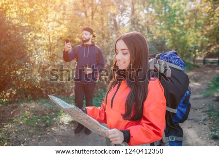 Picture of confident and cheerful young woman with bakcpack. She holds map and looks at it. Young man behind try to catch signal on phone. He holds it up.