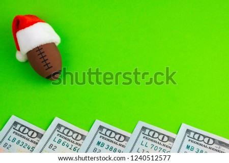 New Year's concept and Christmas sports betting to win or lose. Souvenir ball for American football or rugby in a red Santa Claus hat. Copy space next to dollars on a green background.