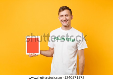 Man in white t-shirt written inscription green title volunteer hold tablet pc computer with blank empty screen isolated on yellow background. Voluntary free assistance help charity grace work concept