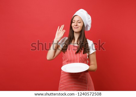 Housewife female chef cook or baker in striped apron white t-shirt, toque chefs hat isolated on red wall background. Woman hold empty blank round plate with place for food. Mock up copy space concept