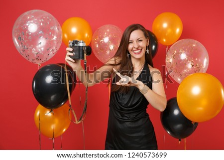 Smiling young girl in black dress doing taking selfie shot, pointing finger on retro vintage photo camera on bright red background air balloons. Happy New Year, birthday mockup holiday party concept