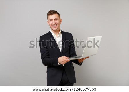 Portrait of laughing young business man in classic black suit, shirt hold laptop pc computer isolated on grey wall background in studio. Achievement career wealth business concept. Mock up copy space