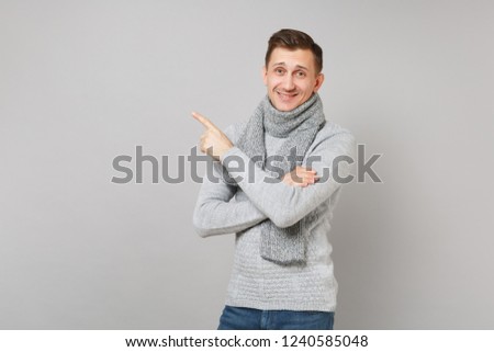 Handsome young man in gray sweater, scarf pointing index finger aside isolated on grey wall background. Healthy fashion, lifestyle people sincere emotions, cold season concept. Mock up copy space