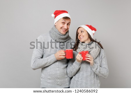 Fun couple girl guy in red Santa Christmas hat gray sweaters scarves hold cups of tea isolated on grey wall background, studio portrait. Happy New Year 2019 holiday party concept. Mock up copy space