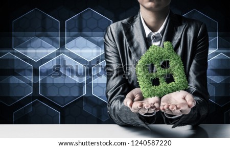 Businessman in suit presenting green plant in form of house symbol with media icons on background.
