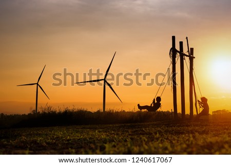 Silhouettes of happiness life of child to playing swing in turbine wind park renewable on sunset. Family Responsibility, Corporate Social Responsibility and sustainable energy concept.