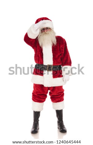 merry saint nick wearing santa costume stands on white background and holds hand on forehead while looking far away, full length picture