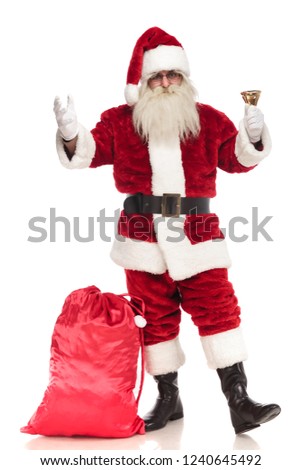 santa claus standing on white background with sack near his leg rings his bell and makes a welcoming gesture, full body picture