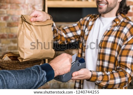 cropped view of smiling seller holding paper bag while customer paying for purchase with credit card