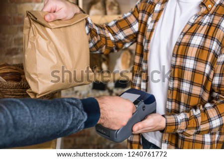 close up of seller holding paper bag while customer paying for purchase with credit card at bakery