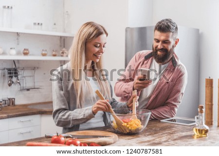 smiling bearded man taking picture of woman cooking salad at kitchen