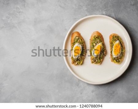 Whole-grain toasts plated avocado with eggs, fresh mini herbs, sunflower seeds served on white craft ceramic plate over grey concrete background, top view, copy space. Diet food concept, gluten-free.