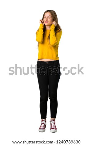 A full-length shot of a young girl with yellow sweater covering ears with hands. Frustrated expression on isolated white background