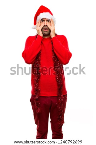 Man with red clothes celebrating the Christmas holidays surprised and covering face with hands while looking through fingers on isolated white background