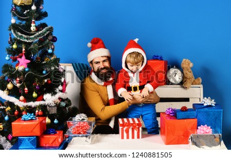 Santa and little assistant among gift boxes near Christmas tree. Christmas family opens presents on blue background. Family holidays concept. Man with beard and smiling face play with son on New Year