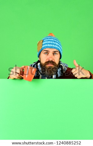 Hipster with beard and concerned face holds tea or coffee cup and shows thumbs up. October beverage idea. Man in hat holds brown cup on green background, copy space. Autumn and cold weather concept