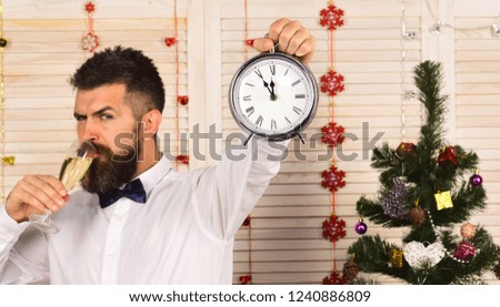 Party man with tricky face drinks holiday beverage. Guy near Christmas tree on wooden wall background. Man with beard holds glass of champagne and alarm clock. Celebration and New Year time concept