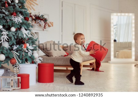 Winter holidays decorations. Warm colors. Beautiful little girl plays with present boxes before a rich decorated Christmas tree in a cozy beige room