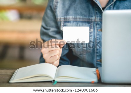 Women's Jean jecket is hand holding white business card for contact works. Blank paper card mock up. Workplace with computer laptop and blank notebook on desk office